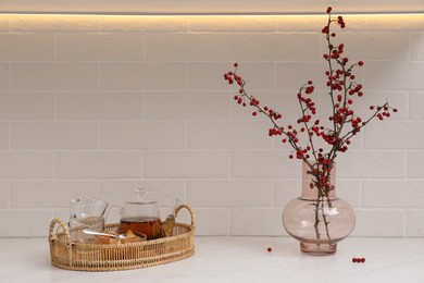 Photo of Hawthorn branches with red berries in vase and aromatic tea on white table indoors