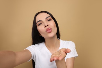 Beautiful young woman taking selfie while blowing kiss on beige background