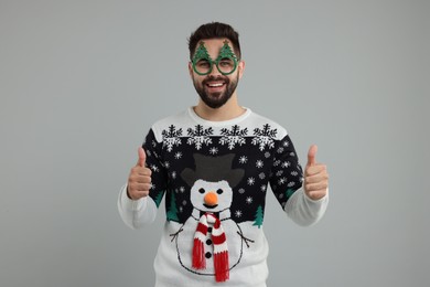 Photo of Happy young man in Christmas sweater and funny glasses showing thumbs up on grey background
