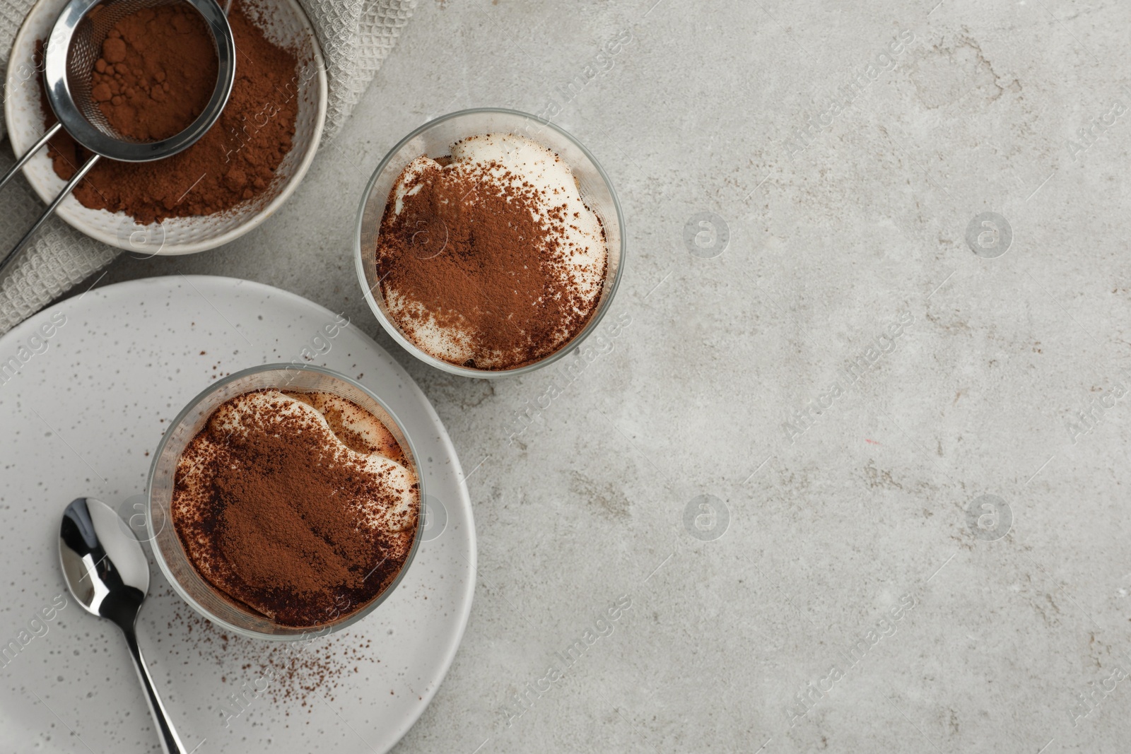 Photo of Delicious tiramisu in glasses, cookies and sieve with cocoa powder on light table, flat lay. Space for text