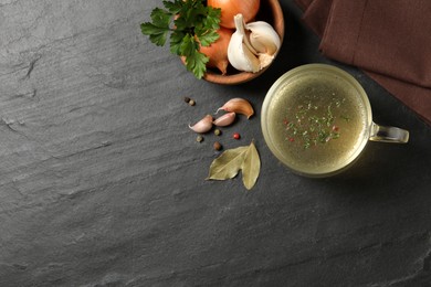 Photo of Hot delicious bouillon in glass cup and ingredients on black table, flat lay. Space for text