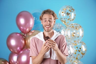 Young man with birthday muffin and air balloons on color background