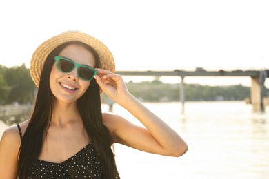 Photo of Beautiful young woman wearing stylish sunglasses near river. Space for text