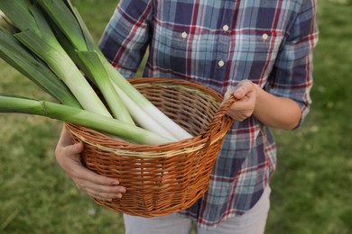 Photo of Woman holding wicker basket with fresh raw leeks outdoors, closeup