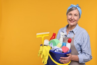 Happy housewife holding bucket with cleaning supplies on orange background, space for text