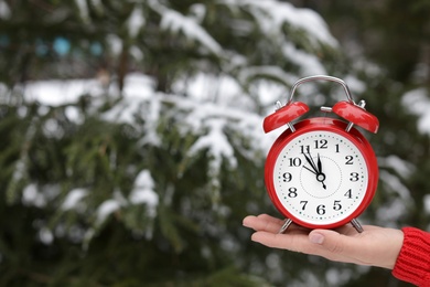 Woman holding red alarm clock near fir tree covered with snow outdoors. Space for text