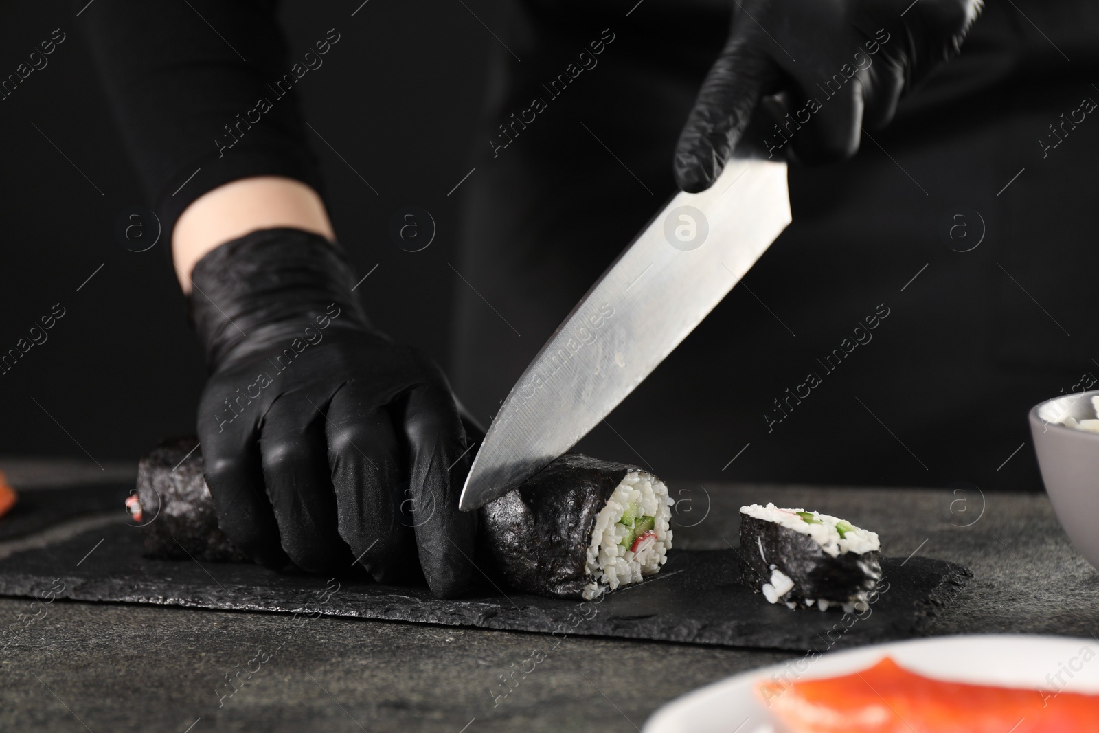 Photo of Chef in gloves cutting sushi roll at dark textured table, closeup