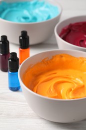 Bowls of different cream with food coloring and bottles of bright liquid on white wooden table, closeup