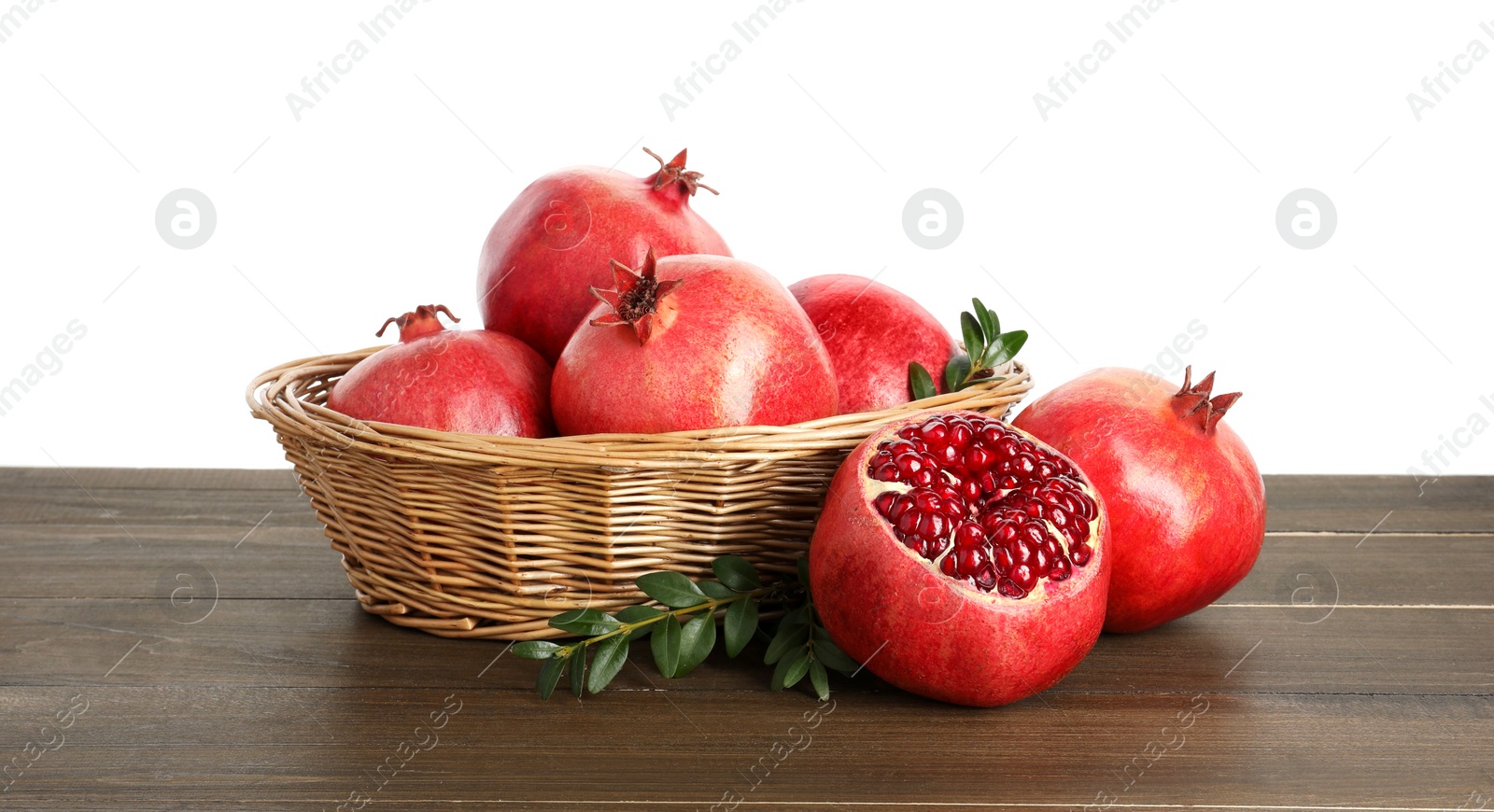 Photo of Fresh pomegranates in wicker basket and green leaves on wooden table against white background