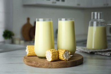 Tasty fresh corn milk in glasses and cobs on white table indoors