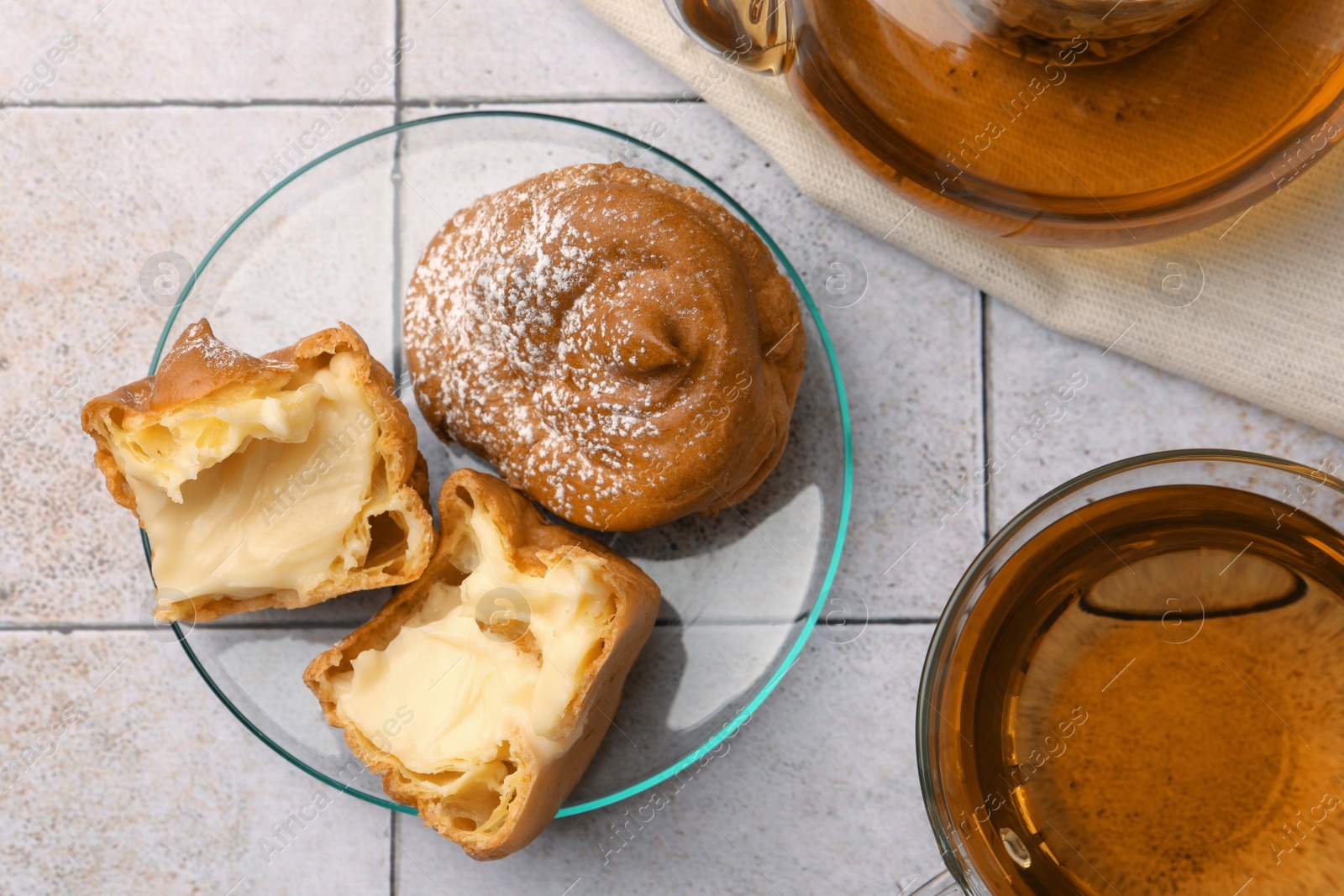 Photo of Delicious profiteroles filled with cream and tea on white tiled table, flat lay