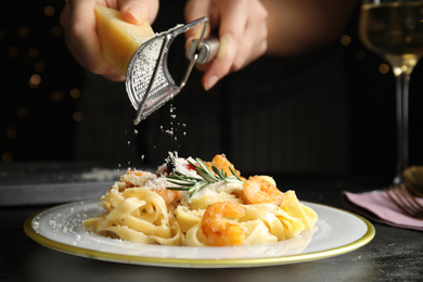 Photo of Woman grating cheese onto pasta with shrimps on black table, closeup