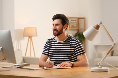 Photo of Home workplace. Happy man taking notes while working with computer at wooden desk in room