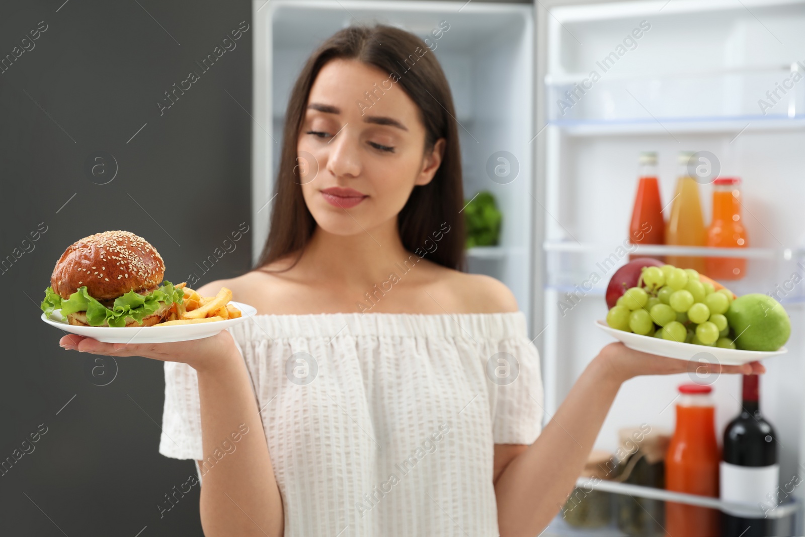 Photo of Woman choosing between fruits and burger with French fries near refrigerator in kitchen