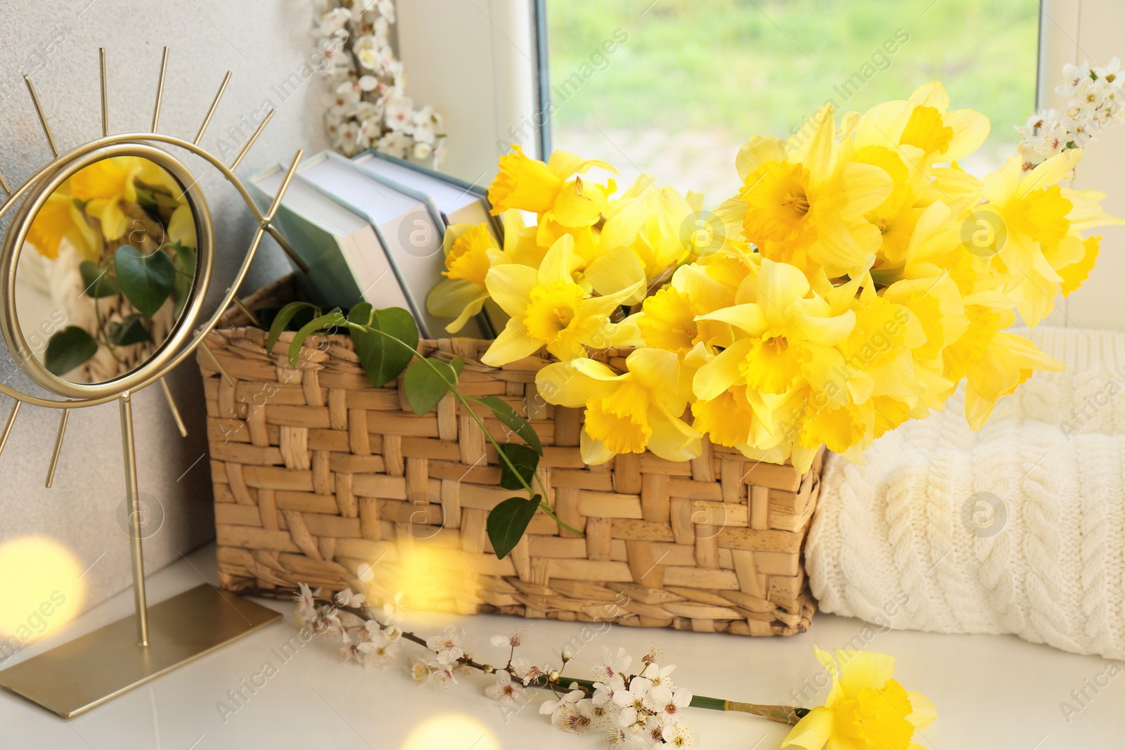Photo of Beautiful yellow daffodils, plum tree branches, wicker basket and mirror on windowsill, closeup. Bokeh effect