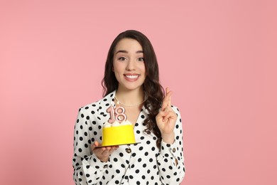 Coming of age party - 18th birthday. Woman holding delicious cake with number shaped candles and crossing fingers on pink background