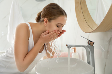 Photo of Young woman washing face with tap water in bathroom
