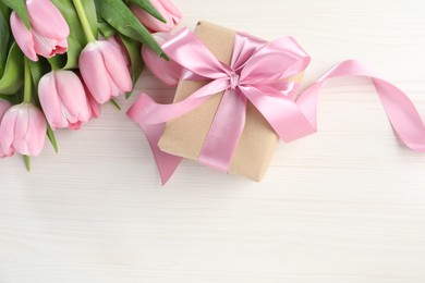 Beautiful gift box with bow and pink tulip flowers on white wooden background, flat lay. Space for text