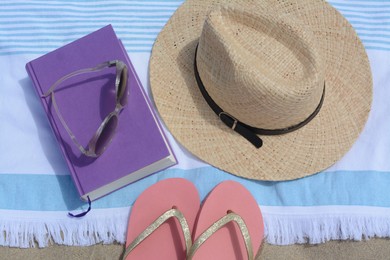 Photo of Beach towel with book, straw hat, sunglasses and flip flops on sand, flat lay