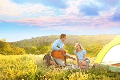 Photo of Young man playing guitar for his girlfriend near camping tent in wilderness