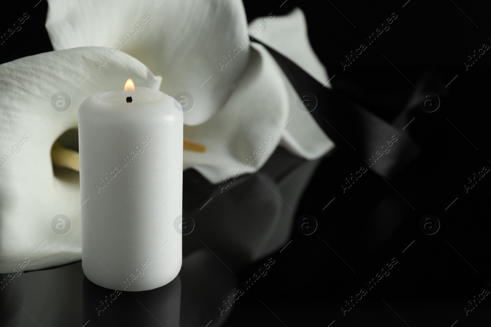 Photo of Burning candle and white calla lily flowers on black mirror surface in darkness, closeup with space for text. Funeral symbol