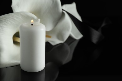 Burning candle and white calla lily flowers on black mirror surface in darkness, closeup with space for text. Funeral symbol