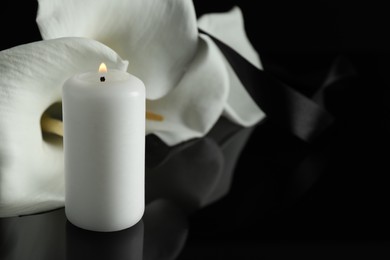 Photo of Burning candle and white calla lily flowers on black mirror surface in darkness, closeup with space for text. Funeral symbol