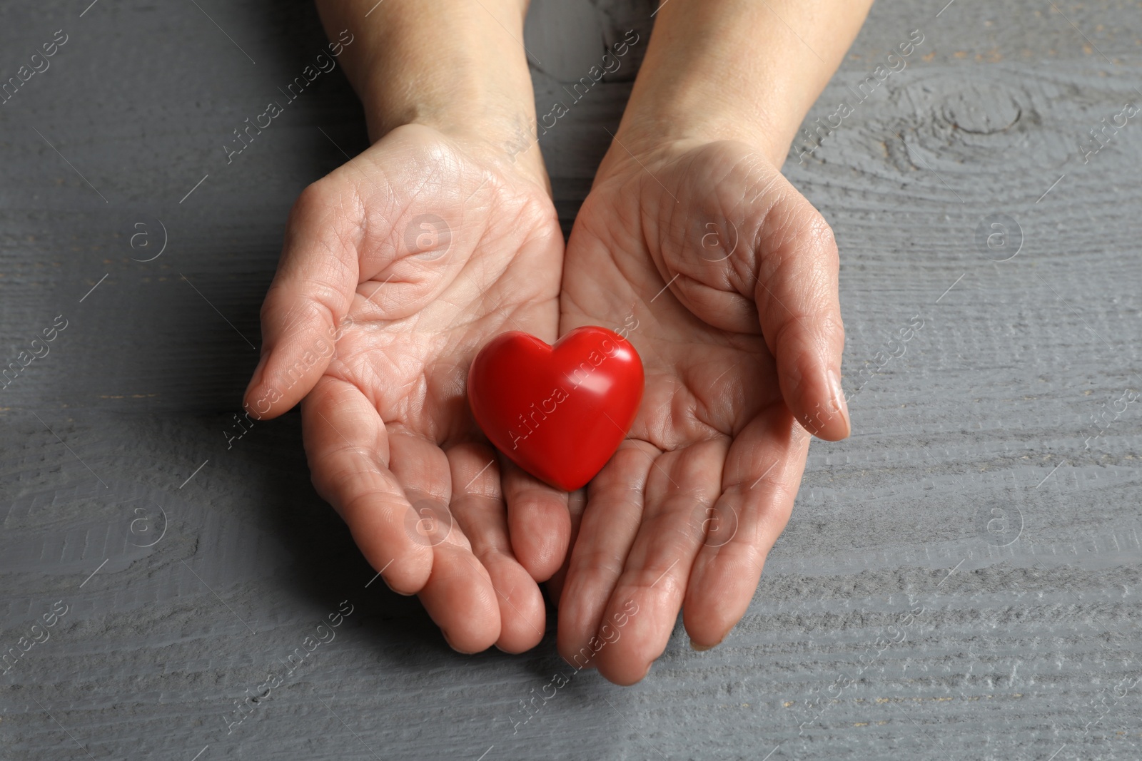 Photo of Elderly woman holding red heart in hands at grey wooden table, closeup