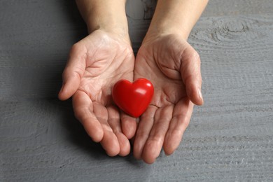 Elderly woman holding red heart in hands at grey wooden table, closeup