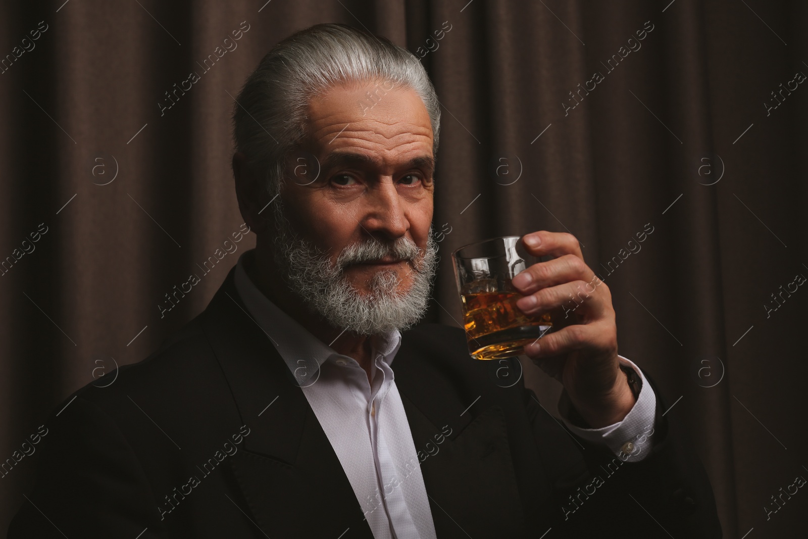 Photo of Senior man in formal suit holding glass of whiskey with ice cubes on brown background