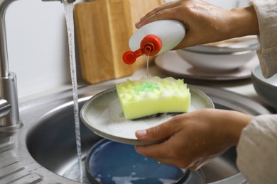 Photo of Woman pouring dishwashing detergent onto plate with sponge near kitchen sink, closeup