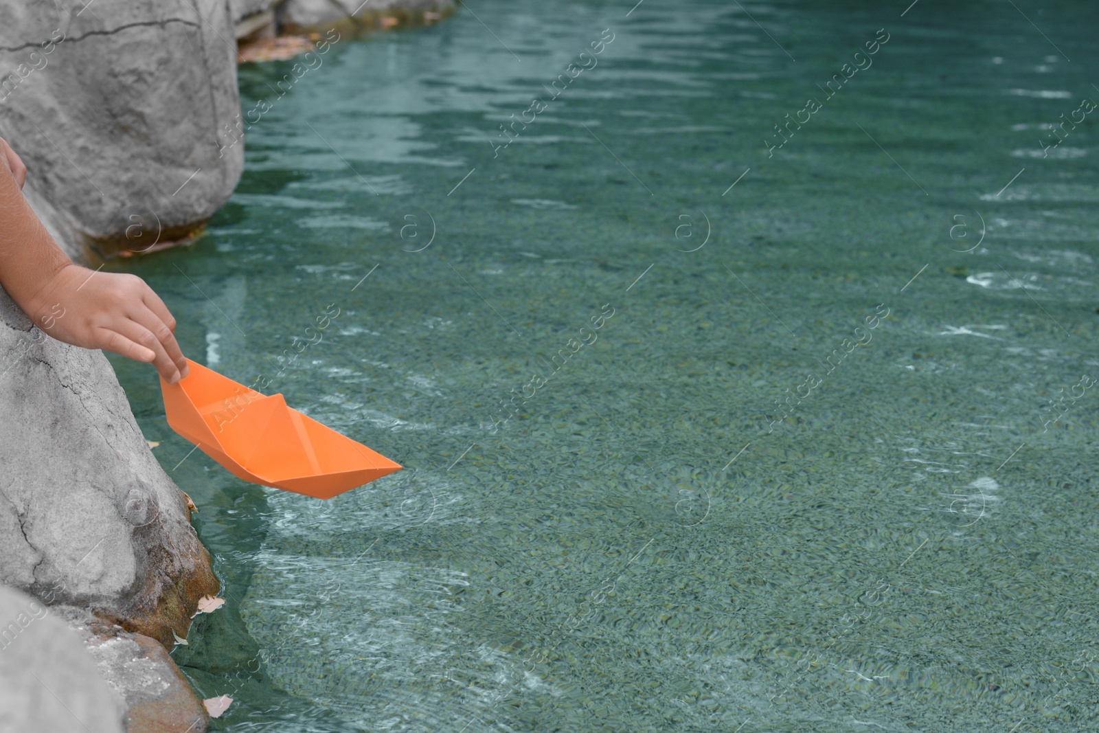 Photo of Kid launching small orange paper boat on water outdoors, closeup. Space for text
