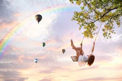 Image of Dream world. Young woman swinging, hot air balloons in sunset sky on background