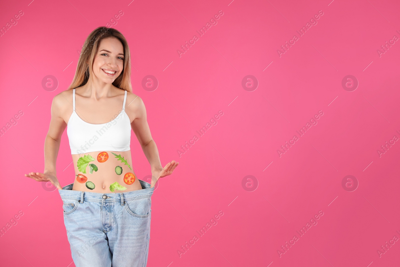 Image of Slim woman in oversized jeans and images of vegetables on her belly against pink background. Healthy eating