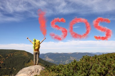 Woman with backpack and word SOS made of color smoke bomb on rocky peak in mountains, back view