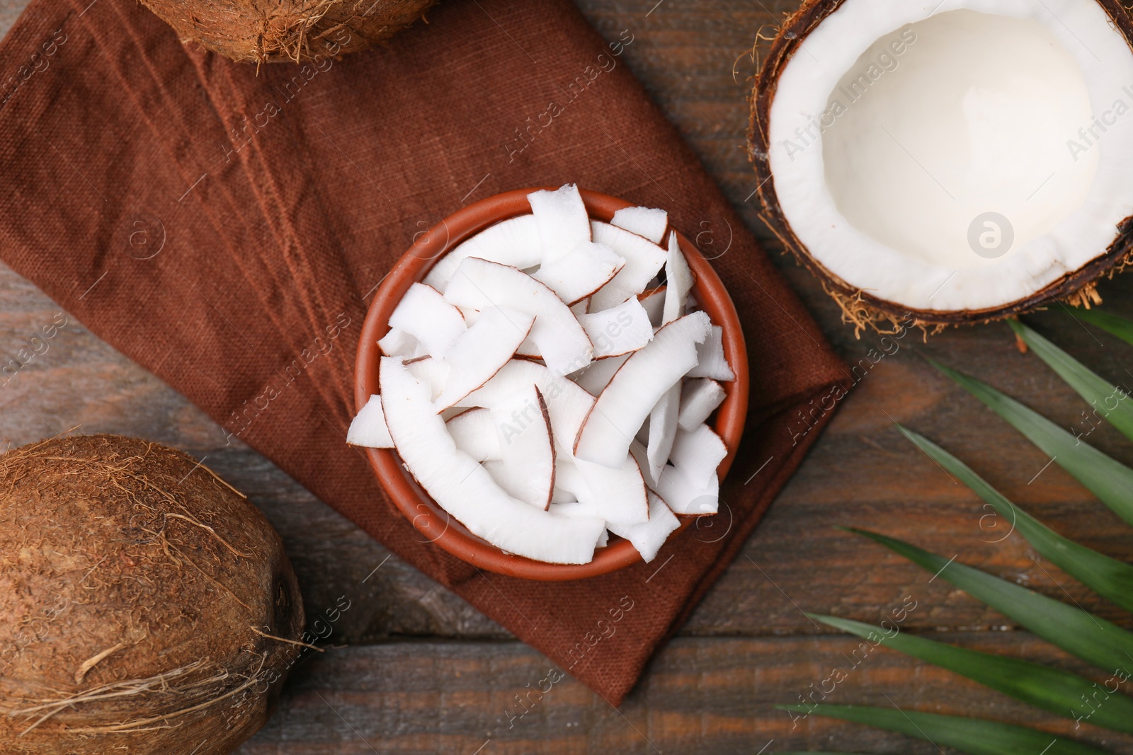 Photo of Coconut pieces in bowl, nuts and palm leaf on wooden table, flat lay