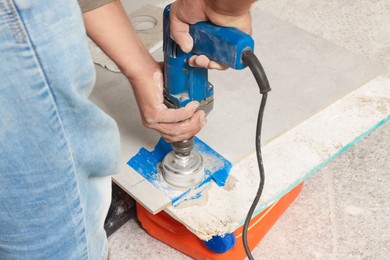 Photo of Worker with electric drill preparing tiles for installation indoors, closeup