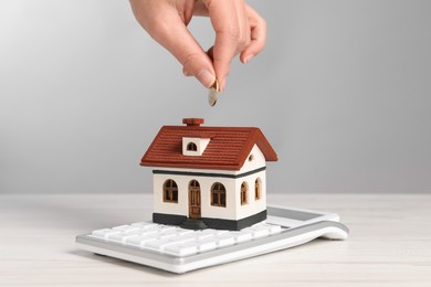 Photo of Mortgage concept. Woman putting coin into house shaped money box at white wooden table, closeup