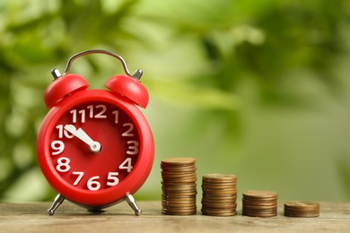 Photo of Red alarm clock and stacked coins on wooden table against blurred background. Money savings