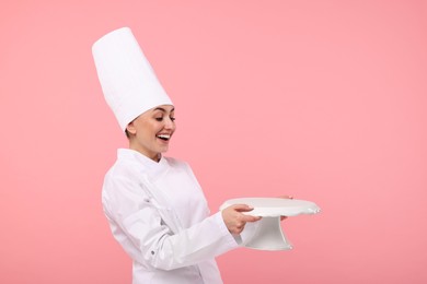 Photo of Happy professional confectioner in uniform holding empty cake stand on pink background. Space for text