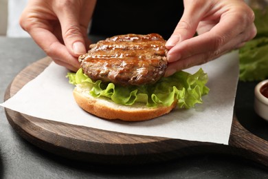 Photo of Woman making tasty hamburger with fried patty, lettuce and bun at table, closeup