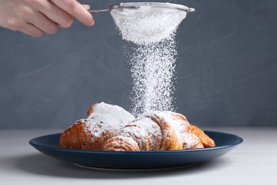 Woman with sieve sprinkling powdered sugar onto croissants at white wooden table, closeup