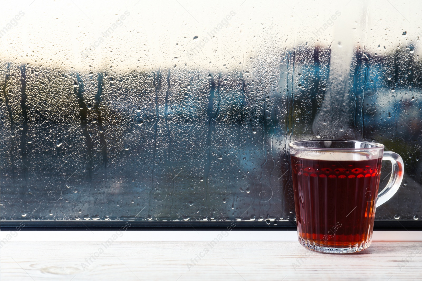 Photo of Glass mug of hot tea on white wooden window sill, space for text. Rainy weather