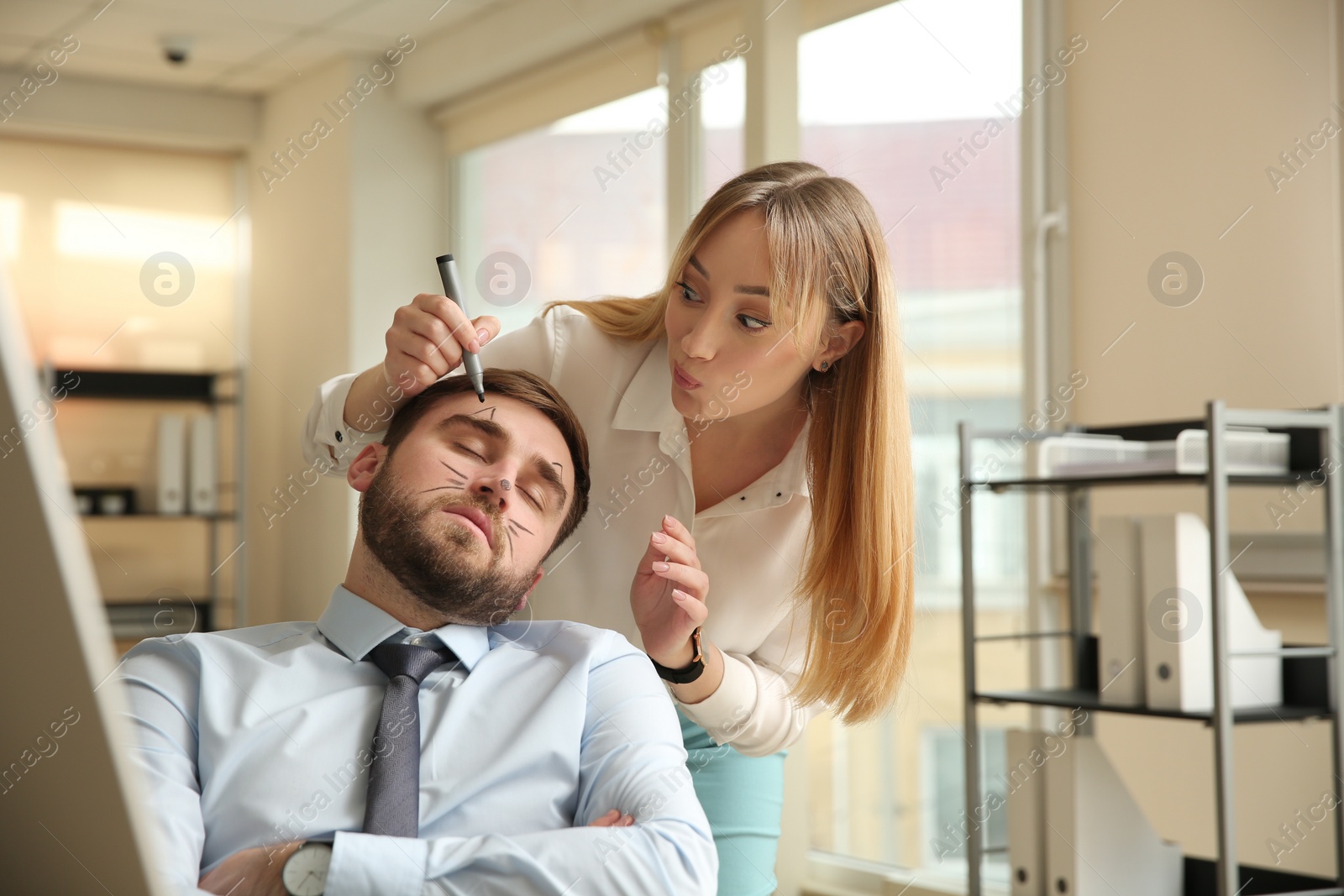 Photo of Young woman drawing on colleague's face while he sleeping in office. Funny joke