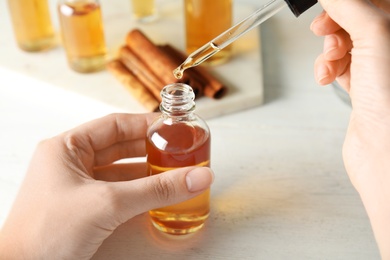 Photo of Woman dripping cinnamon essential oil from pipette into bottle at table, closeup