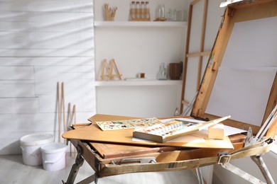 Photo of Foldable wooden easel with empty canvas and supplies in studio. Artist's workplace