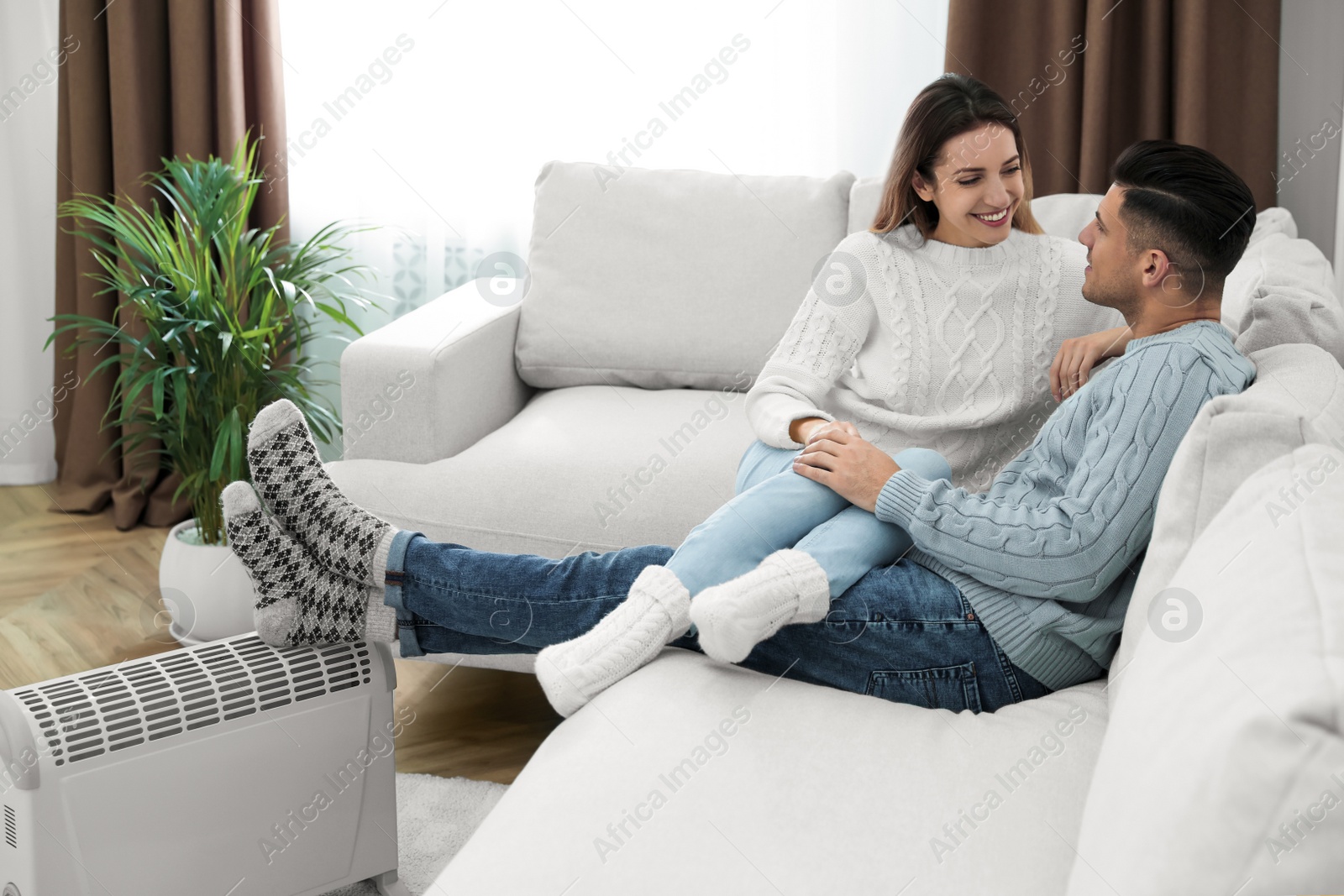 Photo of Happy couple sitting on sofa near electric heater at home