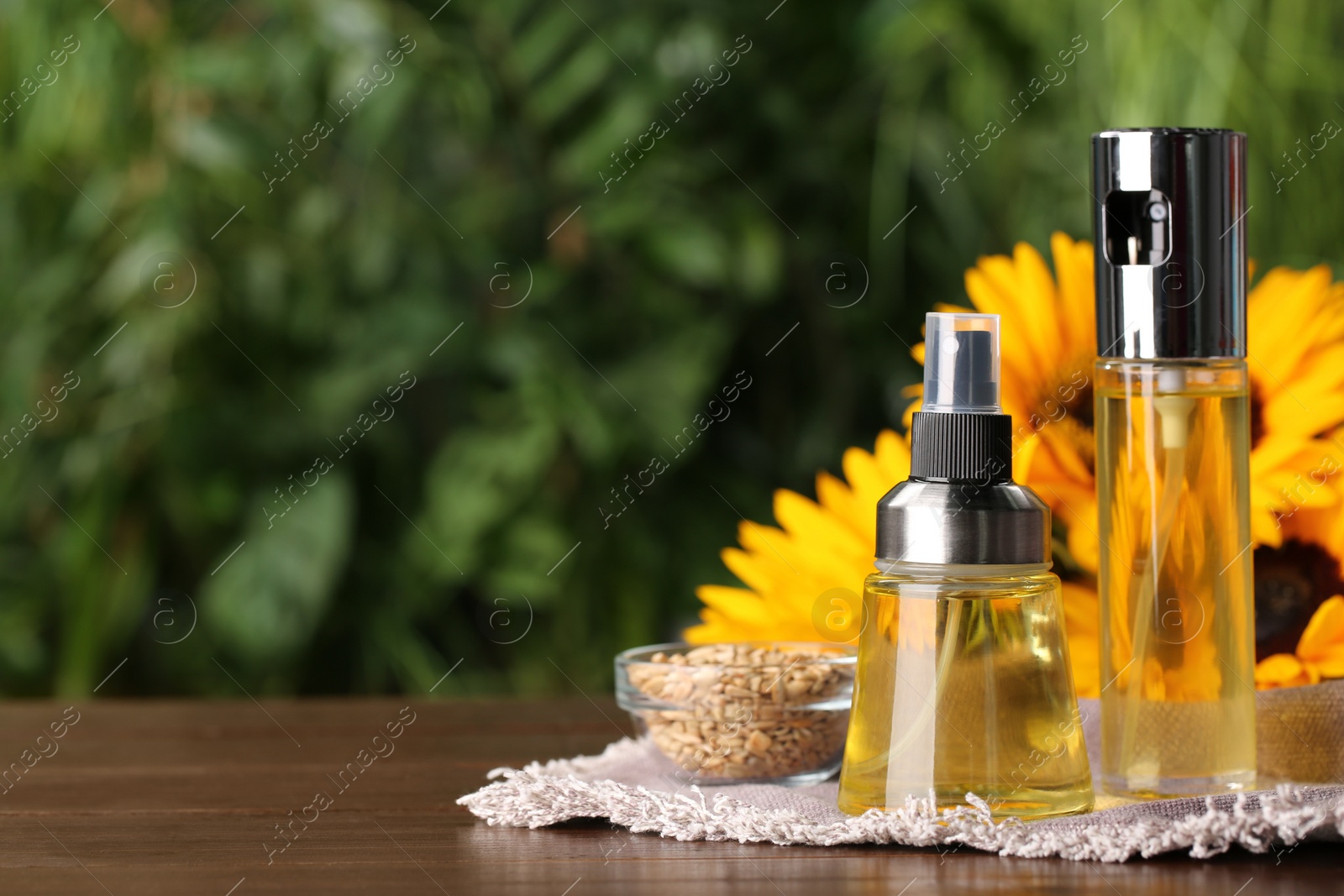 Photo of Spray bottles with cooking oil near sunflower seeds and flowers on wooden table against blurred green background. Space for text