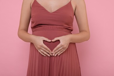 Photo of Pregnant woman in dress making heart with hands on pink background, closeup