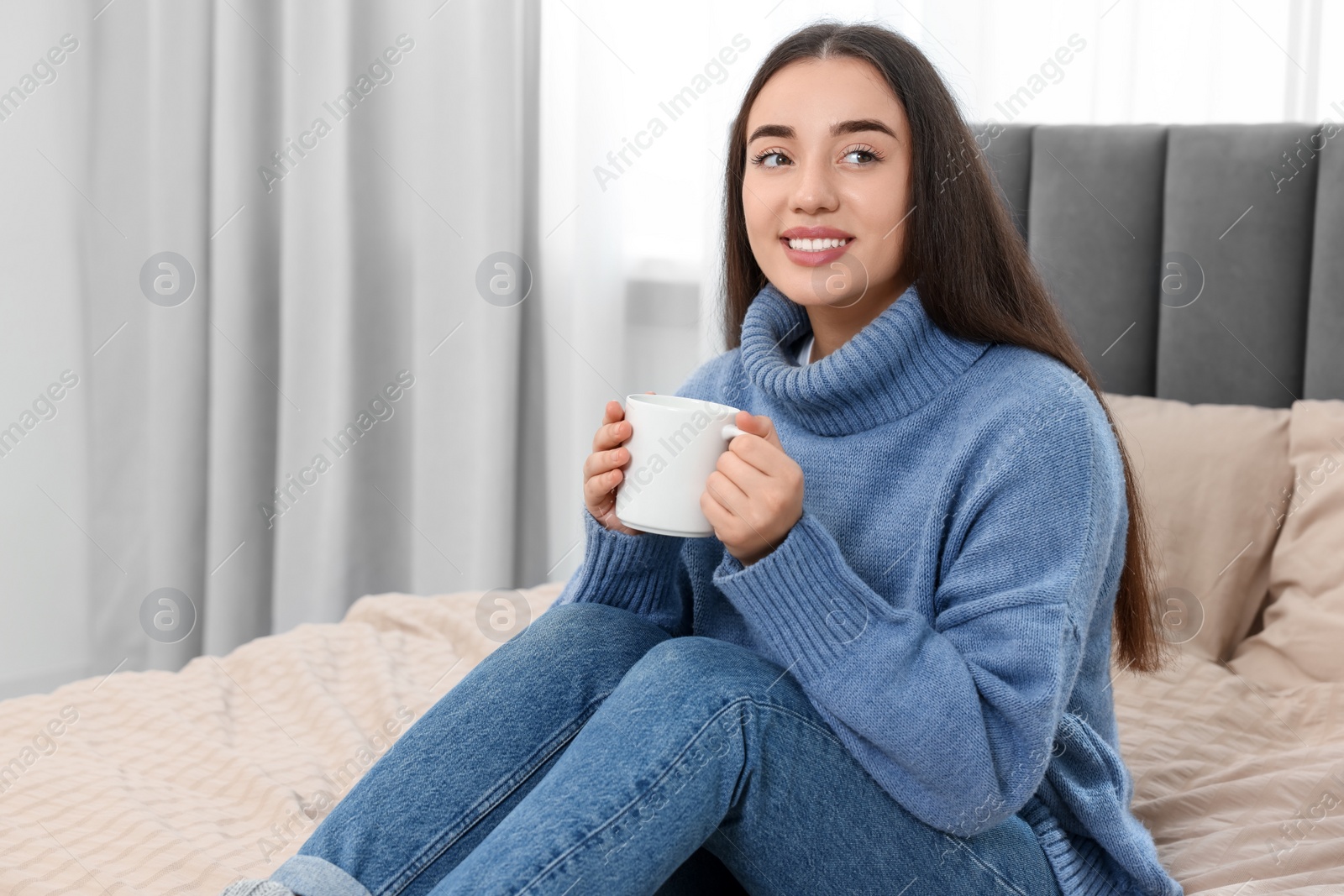 Photo of Happy young woman holding white ceramic mug on bed at home